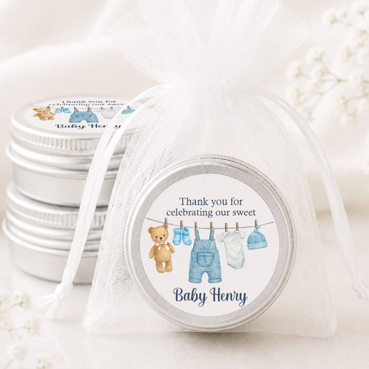 A round baby shower favor tin sealed in a sheer white organza drawstring bag, with three matching favor tins stacked behind it. The round lid features a watercolor illustration of a teddy bear, blue baby clothes on a clothesline, and the text 'Thank you for celebrating our sweet Baby Henry'.