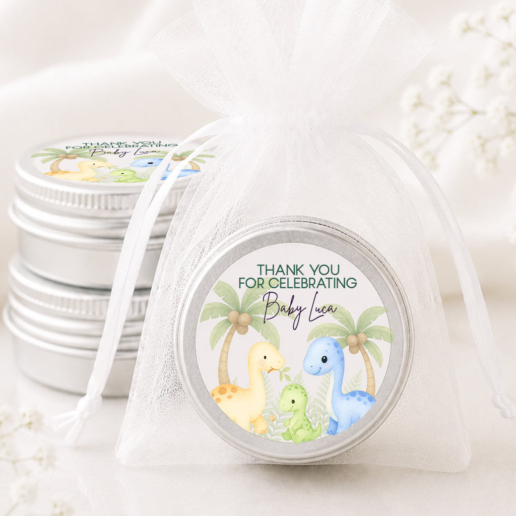 A small round silver tin wrapped in a sheer white organza drawstring bag, with a dinosaur-themed 'Thank You for Celebrating Baby Leo' label on top. More matching silver tins are stacked in the background on a soft white fabric surface.