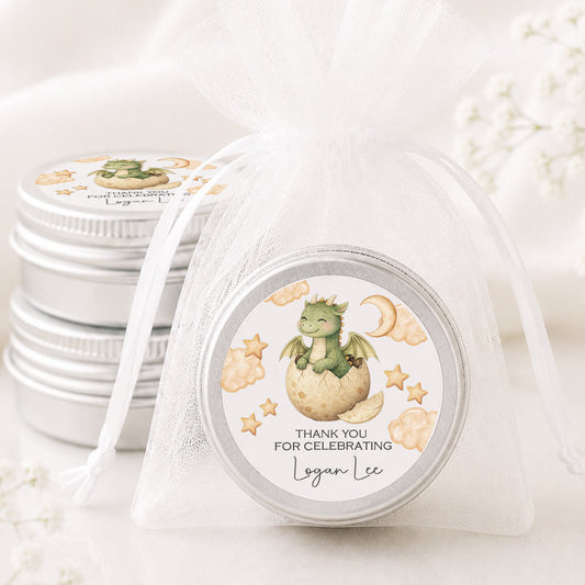 A small round silver tin wrapped in a sheer white organza bag, with a printed circular label showing a green dragon hatching from an egg under a crescent moon and stars. More matching silver tins are stacked in the background on a soft white fabric surface.