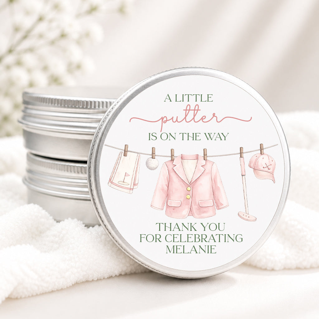 A round silver metal tin with a printed lid design featuring a pink baby outfit hanging on a clothesline, with the text 'A LITTLE putter IS ON THE WAY' and 'THANK YOU FOR CELEBRATING MELANIE.' The tin is shown on soft white fabric with blurred white flowers in the background.