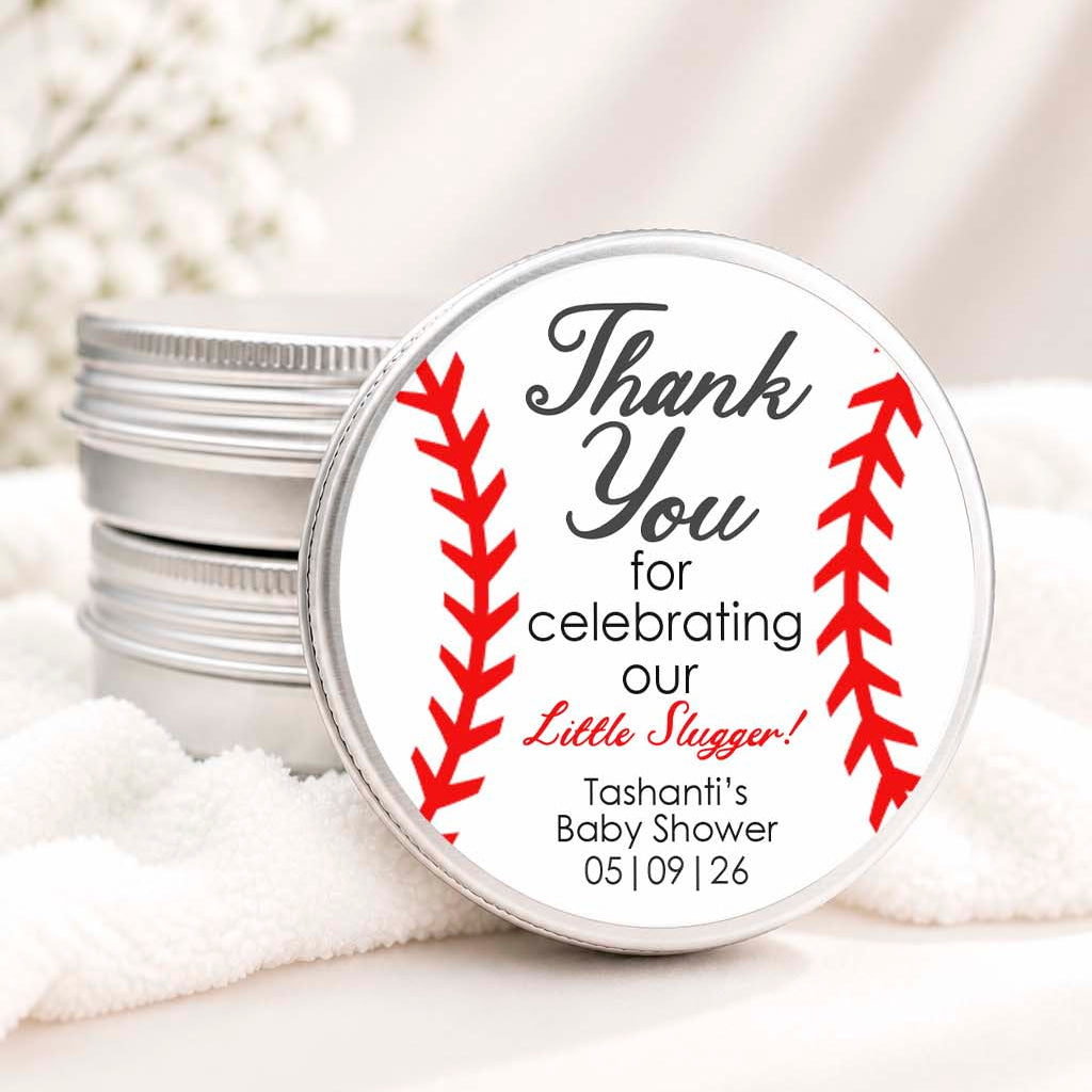 A round silver tin lid with a baseball-themed 'Thank You' baby shower favor label, shown resting on white towels next to stacked silver tins.