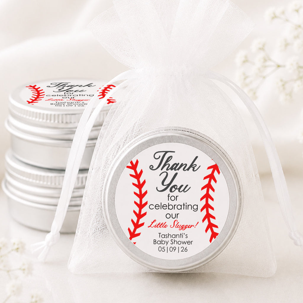 A small round silver tin wrapped in a sheer white organza bag, with a printed round label that reads 'Thank You for celebrating our Little Bunnies' with red baseball-stitch graphics. More matching silver tins are stacked in the background on a soft white fabric surface.