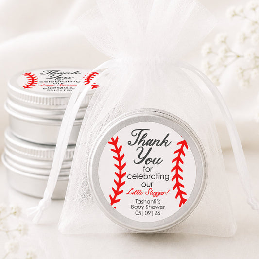 A small round silver tin wrapped in a sheer white organza bag, with a printed round label that reads 'Thank You for celebrating our Little Bunnies' with red baseball-stitch graphics. More matching silver tins are stacked in the background on a soft white fabric surface.