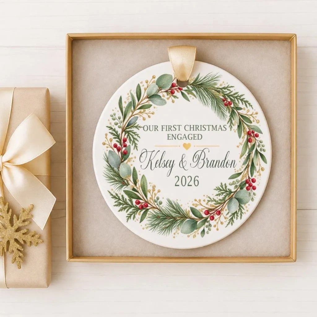 A white ornament with a gold ribbon, featuring a wreath design with greenery, berries, and a heart, is displayed in a gold box.