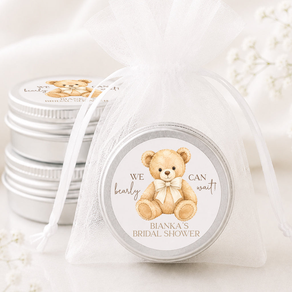 A round silver tin gift box with a personalized teddy bear label, wrapped in a sheer white organza drawstring bag, with two matching silver tins stacked in the background on a white fabric surface.