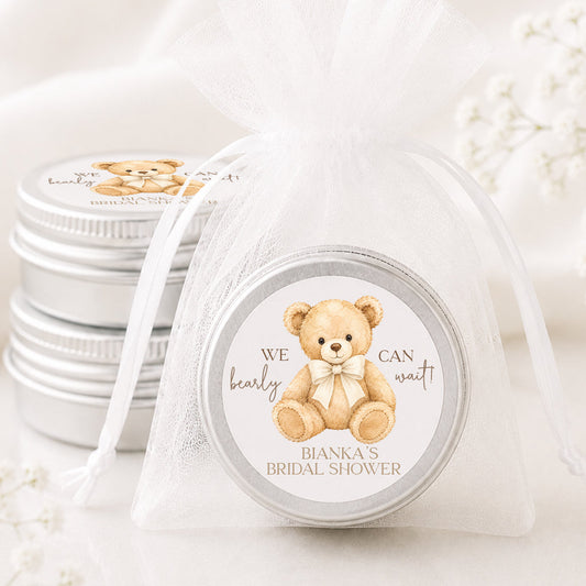 A round silver tin gift box with a personalized teddy bear label, wrapped in a sheer white organza drawstring bag, with two matching silver tins stacked in the background on a white fabric surface.