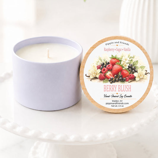 A soy jar candle next to its round lid, which features a berry-themed illustration and reads 'Berry Blush Hand Scented Soy Candle' by peppermint and friends. The candle and lid are displayed on a white ceramic pedestal with a soft-focus background of green leaves and white flowers.