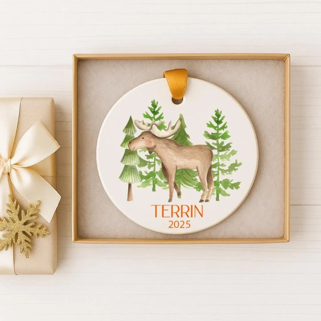 A decorative ornament featuring a moose standing in a forest setting, with the text "TERRIN 2025" printed on it. The ornament is displayed in a gold box, and there is a gift box with a gold ribbon next to it.