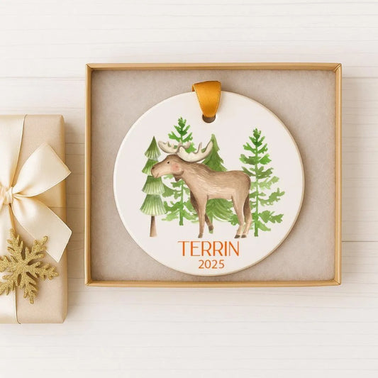 A decorative ornament featuring a moose standing in a forest setting, with the text "TERRIN 2025" printed on it. The ornament is displayed in a gold box, and there is a gift box with a gold ribbon next to it.