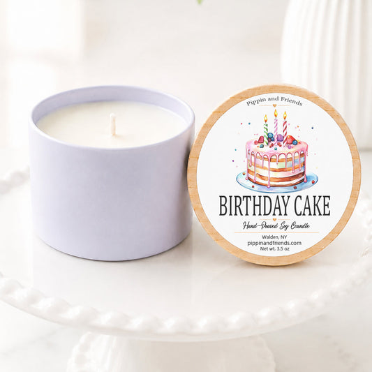 A soy candle in a matte cup-style container sits beside its round lid on a white pedestal. The lid features a printed illustration of a birthday cake and the text 'BIRTHDAY CAKE' with smaller text above reading 'Pippin and Friends'.