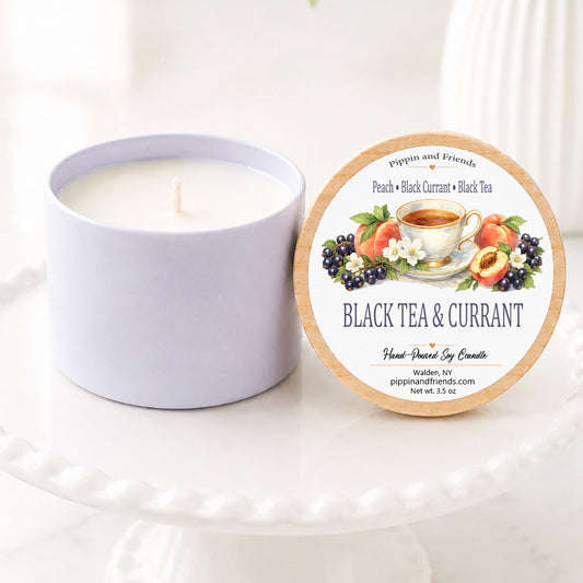 A soy candle in a matte container sits next to its round illustrated label on a white pedestal stand. The label reads 'Black Tea & Currant' and features illustrations of a tea cup with strawberries, blueberries, and peaches.