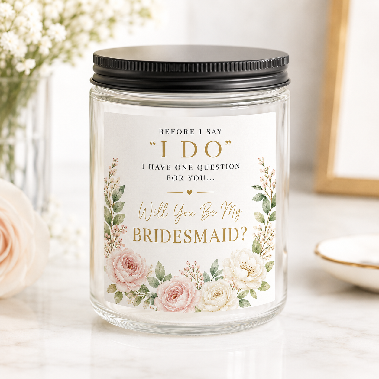 Elegant bridesmaid proposal candle in a clear glass jar with black lid featuring blush floral artwork and the message "Will You Be My Bridesmaid?" surrounded by soft pink and ivory flowers.