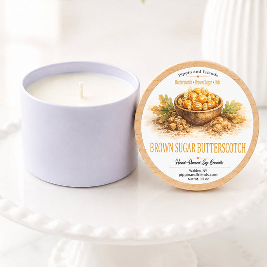 A soy candle jar sits beside its round lid on a white pedestal. The lid features a natural wood border and an illustration of brown sugar in a bowl, with the text 'BROWN SUGAR BUTTERSCOTCH' and the brand 'pippin and friends'.
