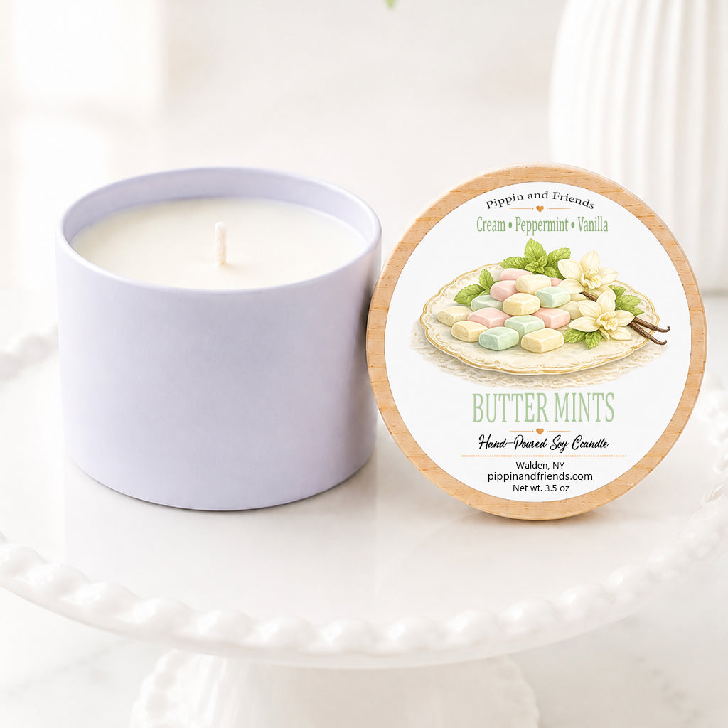 Mint scented hand-poured soy candle in a matte white vessel with natural wooden lid displayed upright beside it, featuring a watercolor plate of butter mints label. The candle sits on a white scalloped pedestal against a bright, airy background with soft white flowers in a vase, creating a clean, cozy woodland-inspired aesthetic.