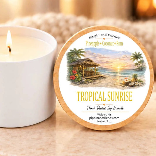 A white candle with a tropical island scene on its label, placed next to a lit candle, on a beige surface.