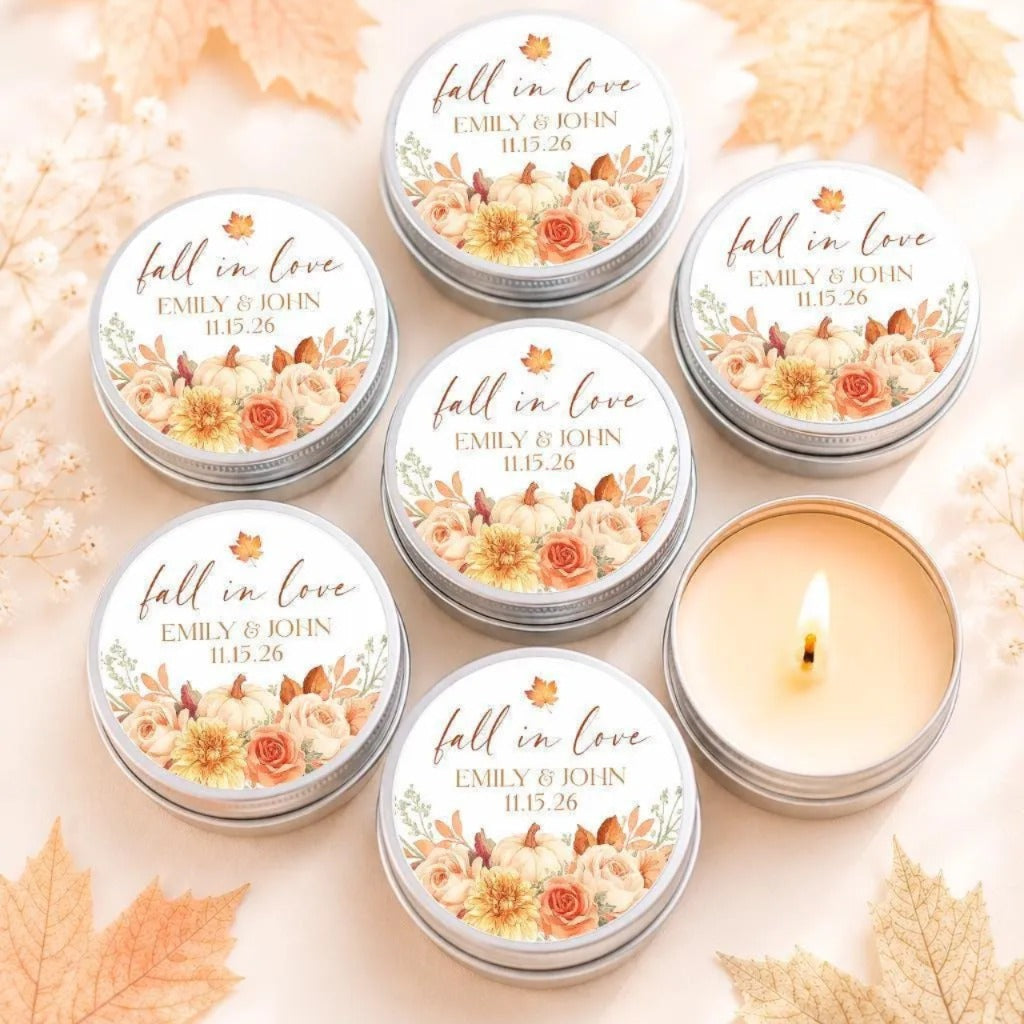 a collection of six round tin candles with floral designs, arranged on a surface with autumn leaves. The candles are labeled with the phrase "fall in love" and the names "EMILY & JOHN" along with the date "11.25.26".