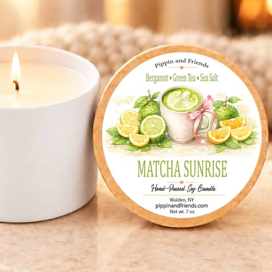 a white candle with a label that reads "MATCHA SUNRISE Hand-Poured Soy Candle" and " Walden, NY pippinandfriends.com net wt 7 oz". The candle is placed next to a white candle holder, both of which are set against a blurred background.