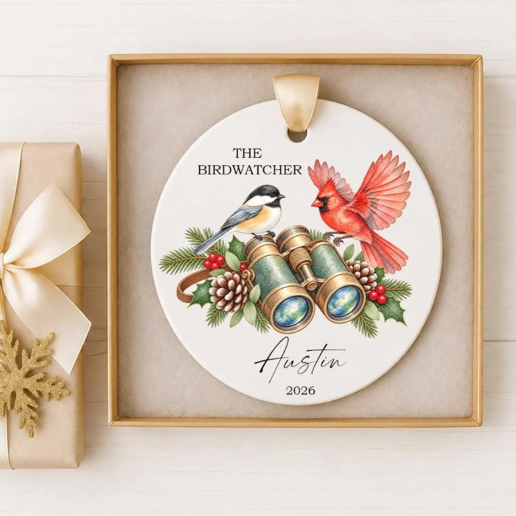 A decorative ornament featuring a birdwatcher theme, with illustrations of a red cardinal and a black and white chickadee. The ornament is displayed in a gold frame, and there is a small gift box next to it.