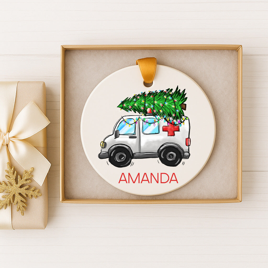 A white Christmas ornament with a Christmas tree on top of a white ambulance, displayed in a gold box.