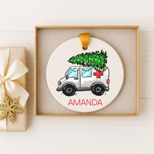 A white Christmas ornament with a Christmas tree on top of a white ambulance, displayed in a gold box.