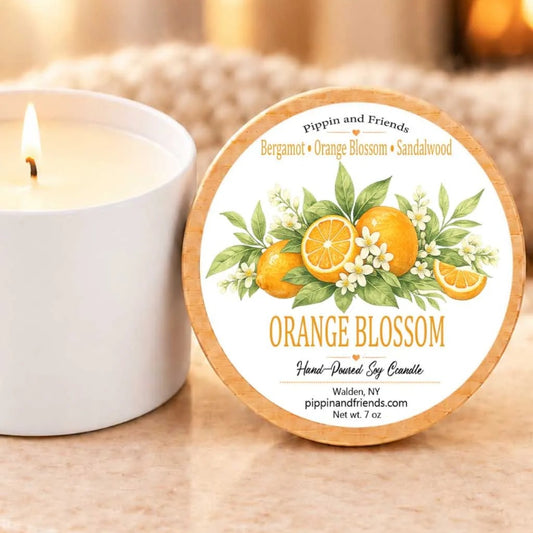 a white candle with a label that reads "Orange Blossom" and "Pippin and Friends Bergamot + Orange Blossom + Sandalwood". The candle is placed next to a white ceramic mug, both resting on a beige surface.