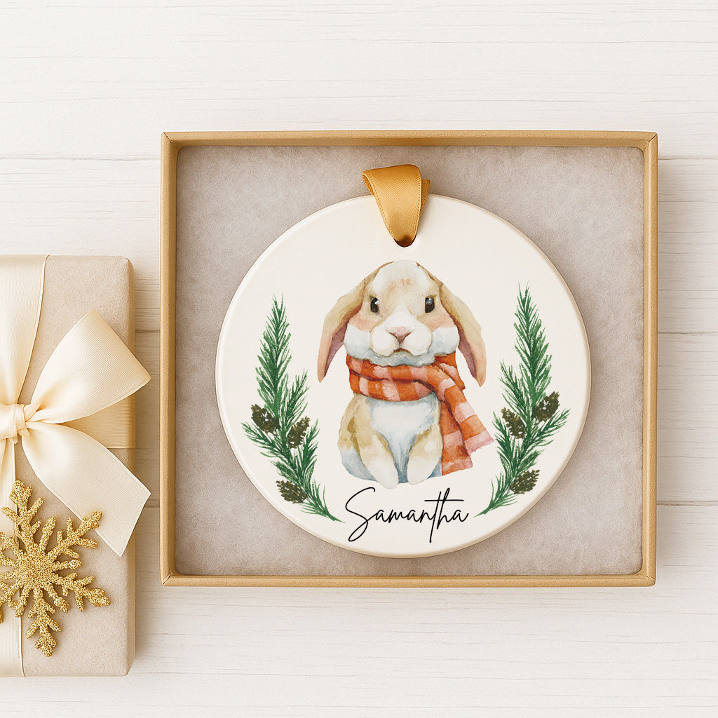 A ceramic ornament featuring a bunny wearing a red scarf and surrounded by pine branches, with the name "Samantha" written on it. The ornament is displayed in a gold frame, and there is a gift box with a gold bow next to it.