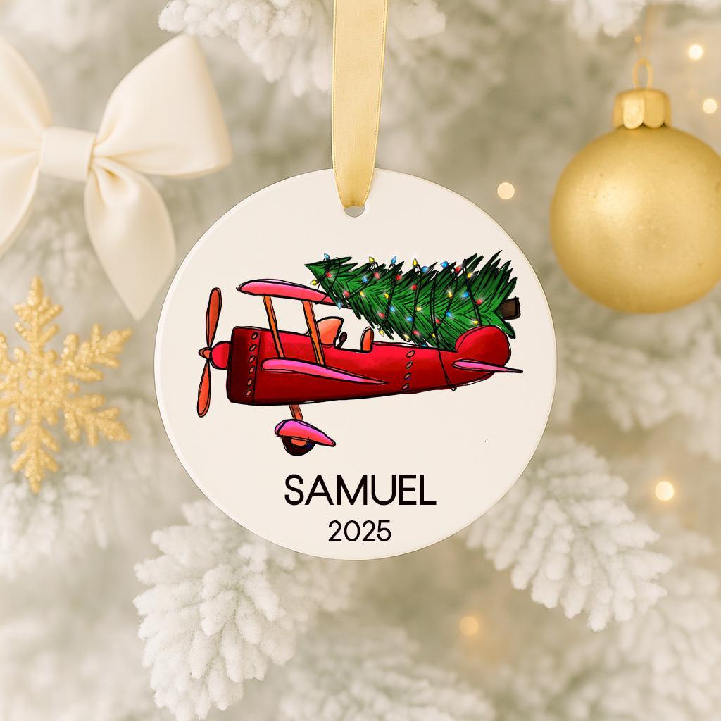 A white ornament with a red biplane carrying a Christmas tree is hanging on a tree.