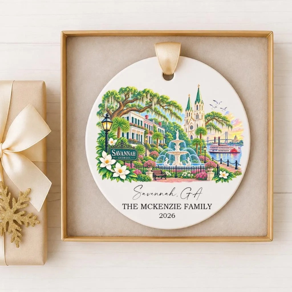 A decorative ornament featuring a scenic view of Savannah, Georgia, with a fountain, buildings, and greenery. The ornament is displayed in a wooden frame and is accompanied by a ribbon.