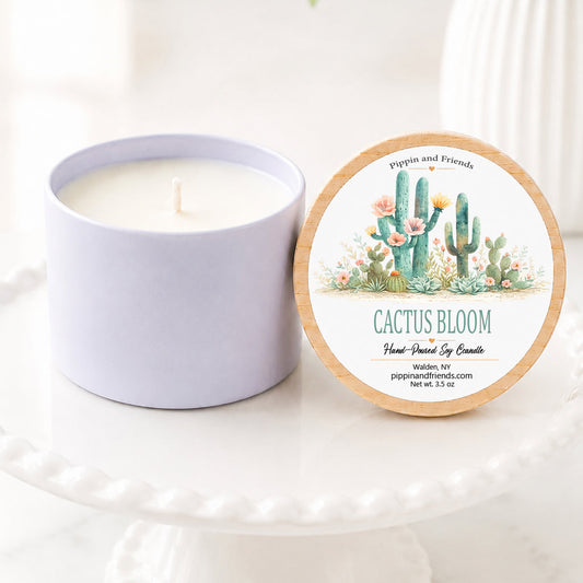 A candle in a white jar sits beside its round lid on a white pedestal. The lid features a cactus illustration and the text 'Cactus Bloom' candle' by Pippin and Friends.