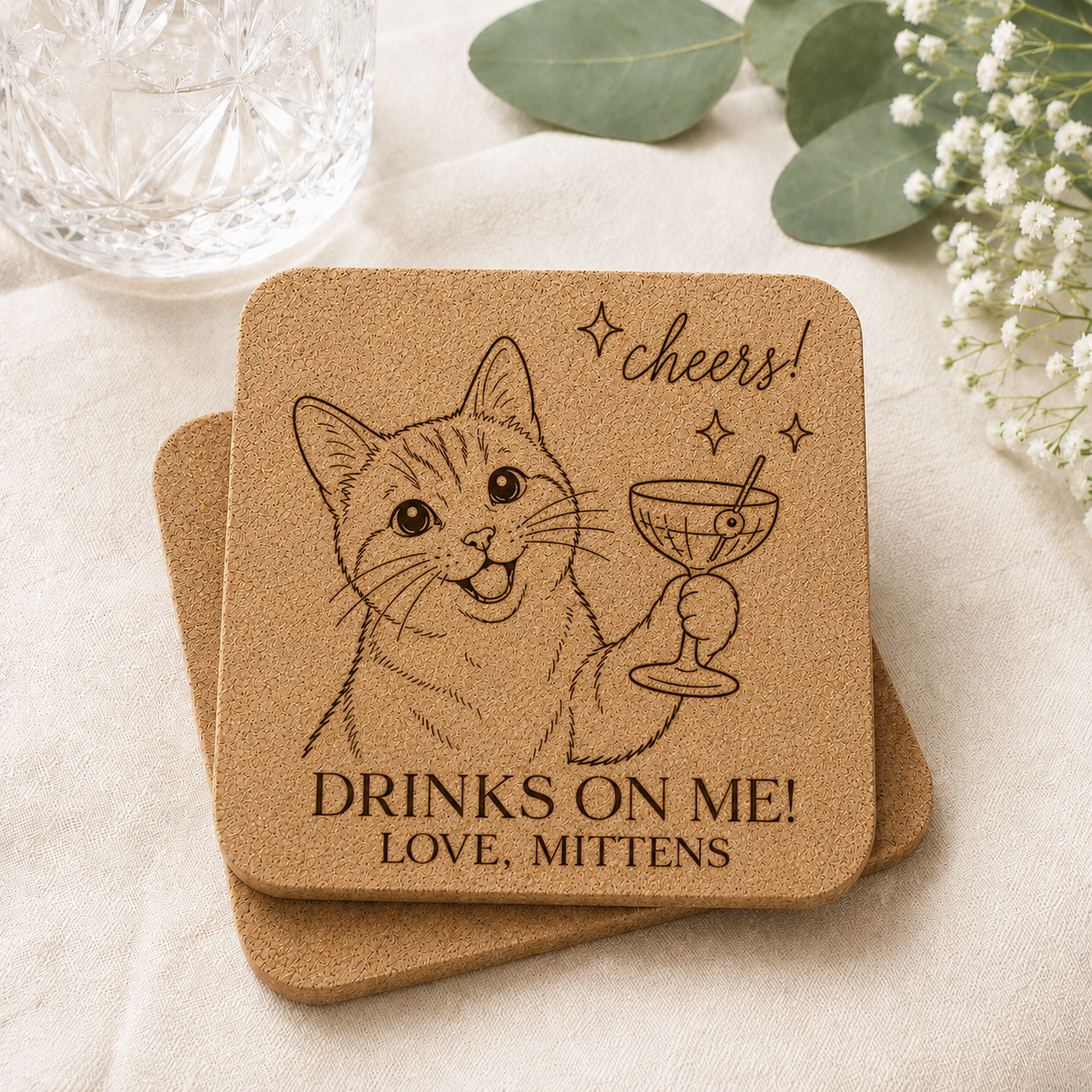 Personalized cork wedding favor coaster engraved with a cat holding a cocktail glass and the phrase "Drinks On Me! Love, Mittens" on a light airy wedding-inspired background.