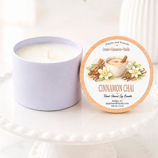 A soy candle jar sits beside its round lid on a white pedestal. The lid features a label for 'Cinnamon Chai' by pippinand friends, decorated with illustrations of cinnamon sticks, a mug of chai, and white flowers.