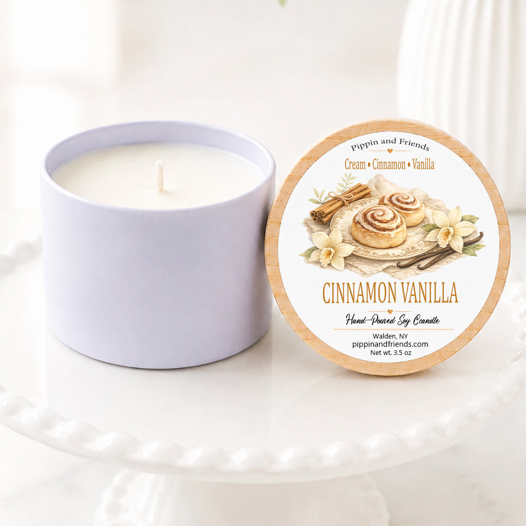 A soy candle jar sits beside its round lid on a white pedestal. The lid features a warm-toned label with an illustration of cinnamon rolls and the text 'Cinnamon Vanilla' and 'pippin and friends'.