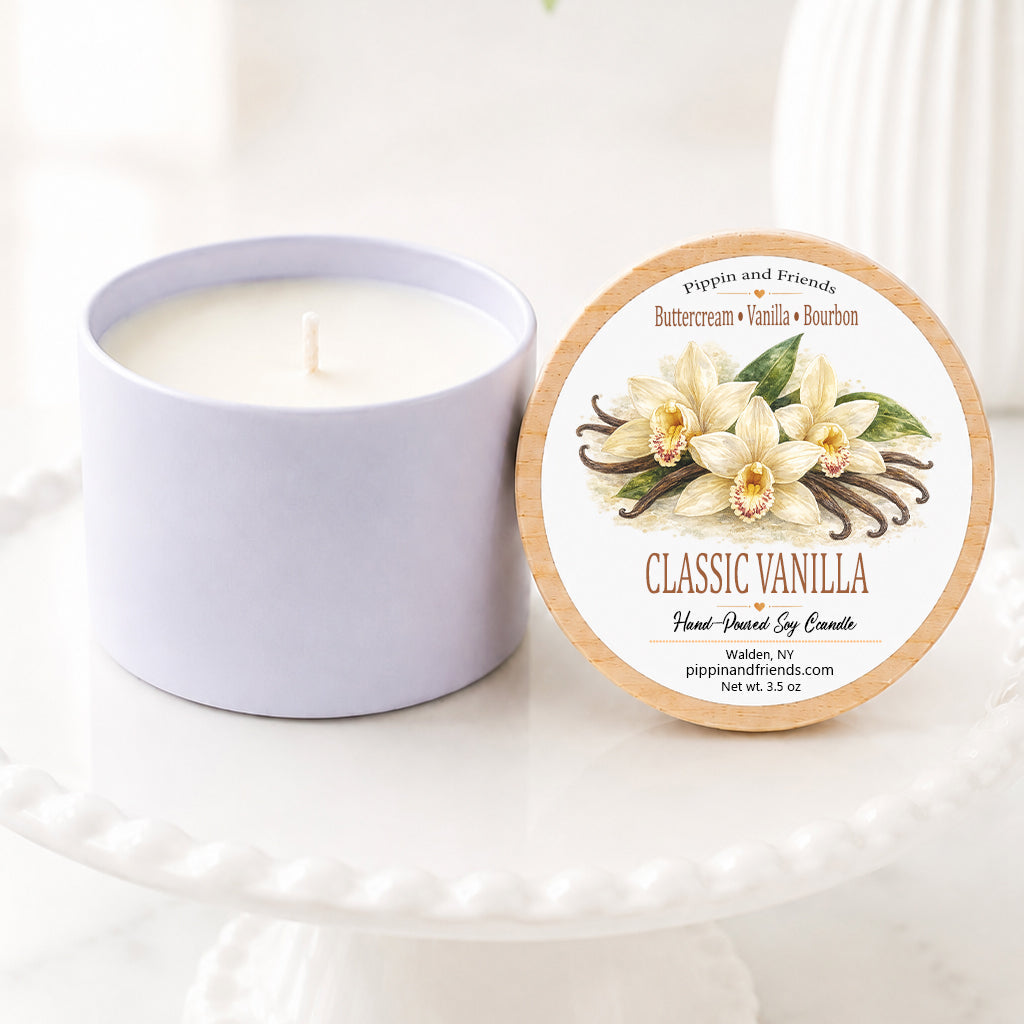 Classic vanilla hand-poured soy candle in a matte white vessel with natural wooden lid displayed upright beside it, featuring a watercolor vanilla flower label. The candle sits on a white scalloped pedestal against a bright, airy background with soft white flowers in a vase, creating a clean, cozy inspired aesthetic.