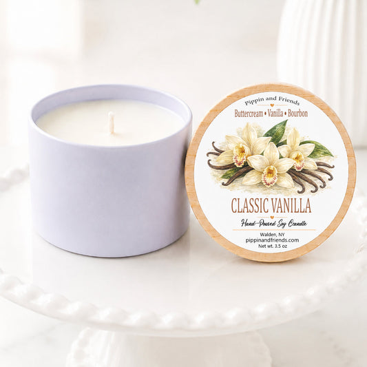 Classic vanilla hand-poured soy candle in a matte white vessel with natural wooden lid displayed upright beside it, featuring a watercolor vanilla flower label. The candle sits on a white scalloped pedestal against a bright, airy background with soft white flowers in a vase, creating a clean, cozy inspired aesthetic.