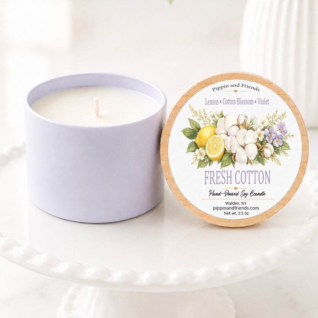 A soy candle in a matte container sits next to its round lid on a white pedestal. The lid features a floral label with the text 'Fresh Cotton' and illustrations of cotton bolls, lemons, and lavender.