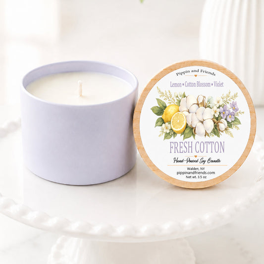 A soy candle in a matte container sits next to its round lid on a white pedestal. The lid features a floral label with the text 'Fresh Cotton' and illustrations of cotton bolls, lemons, and lavender.