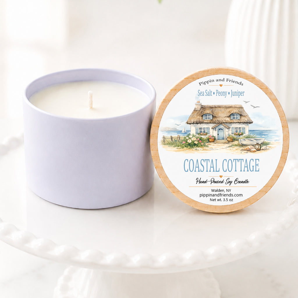 A soy candle in a matte cylinder container sits next to its round lid on a white pedestal. The lid features a wood-grain border and an illustration of a coastal cottage with the text 'COASTAL COTTAGE' and 'pippin and friends'.