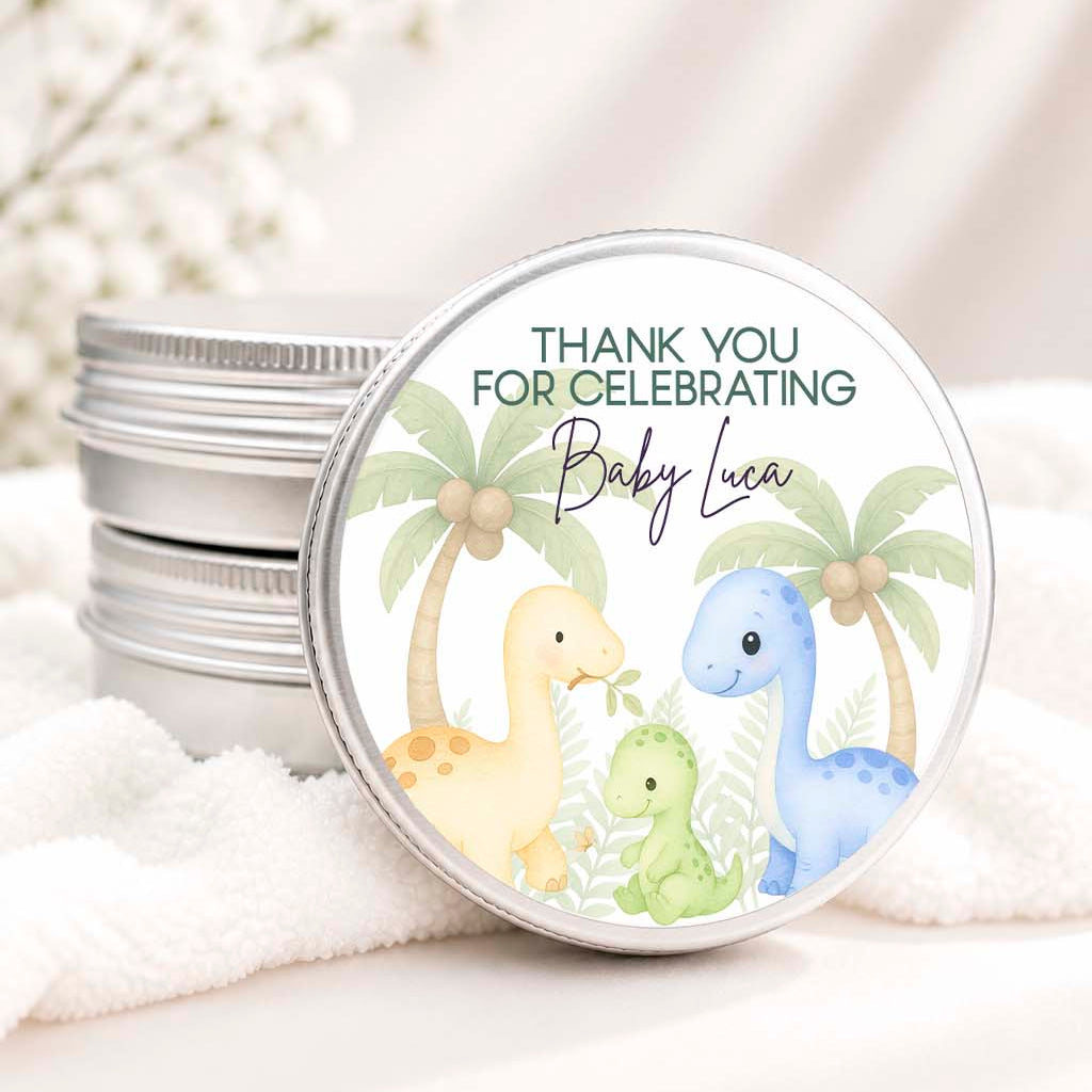 A stack of round silver metal gift tins sits on white fabric, with one tin leaning forward to show a printed lid design. The lid features a watercolor-style illustration of three pastel dinosaurs in yellow, green, and blue with palm trees, plus the text 'THANK YOU FOR CELEBRATING Baby Luca'.