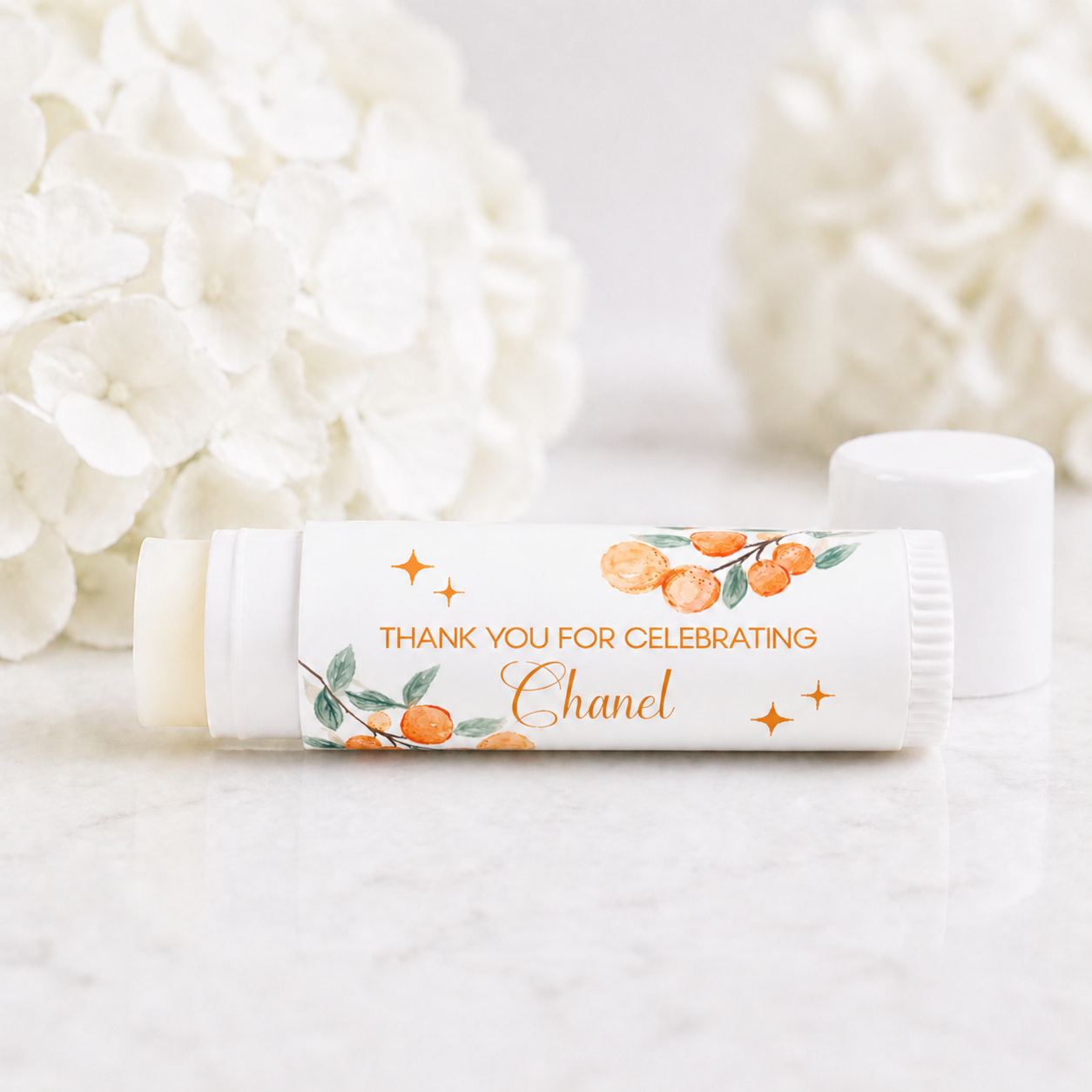 Citrus Cutie baby shower lip balm favor with personalized orange watercolor label reading thank you for celebrating Chanel on white tube with soft floral background