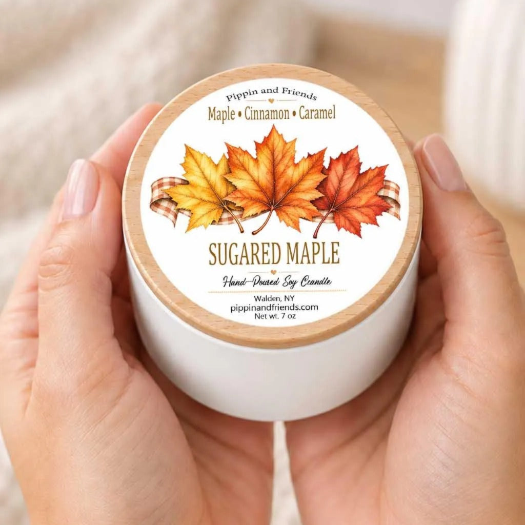 A person's hands holding a white container with a label featuring a design of autumn leaves and the text "SUGARED MAPLE Hand Picked Soy Candle" printed on it.