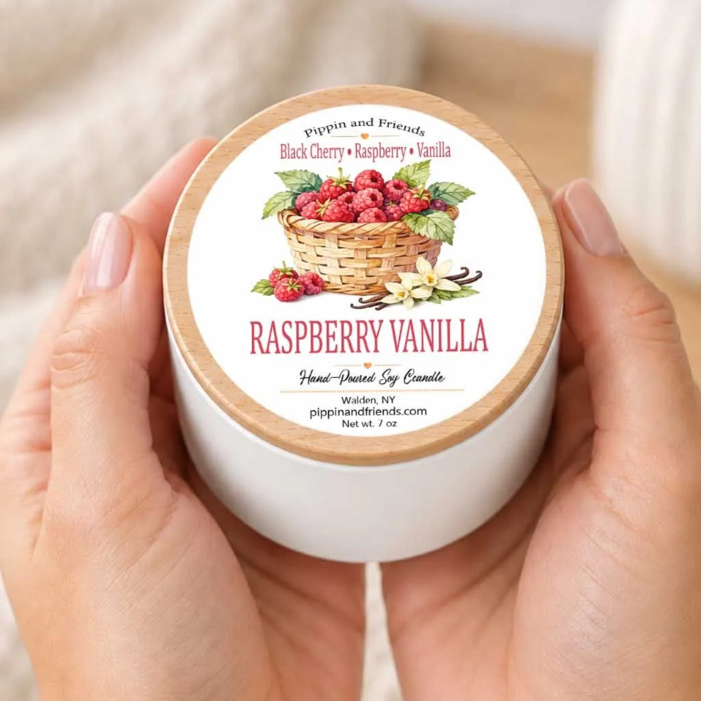 A person's hands holding a white container with a label that features an illustration of a basket filled with raspberries and a label that reads "RASPBERRY VANILLA Hand-poured by Candela".