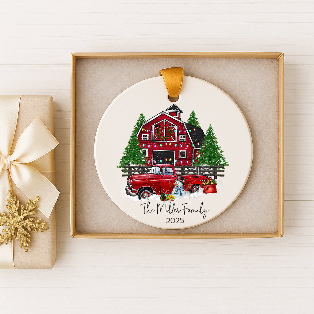 A white ceramic ornament with a red barn and Christmas trees, featuring a red truck and a snowman, is displayed in a gold frame.