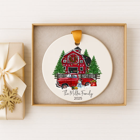 A white ceramic ornament with a red barn and Christmas trees, featuring a red truck and a snowman, is displayed in a gold frame.