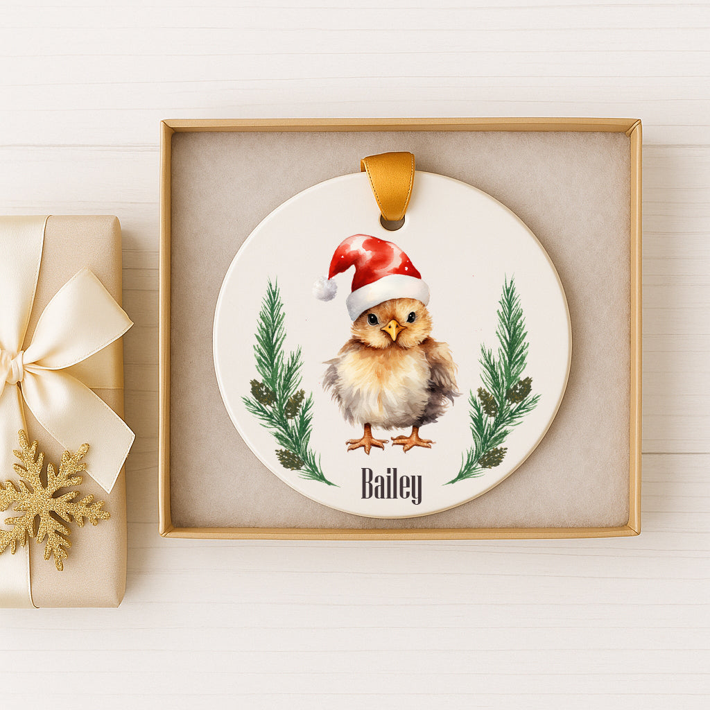 A ceramic ornament featuring a small brown bird wearing a Santa hat, surrounded by pine branches and greenery. The ornament is displayed in a gold box, and there is a gift box with a gold ribbon next to it.