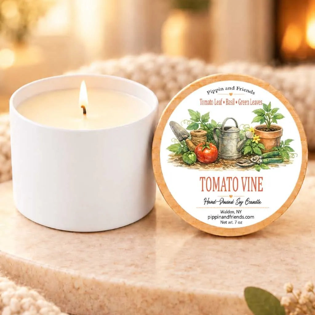 a white candle with a label that reads "Tomato Vine" and "Pippin and Friends" on a wooden surface, with a blurred background that includes a fireplace.