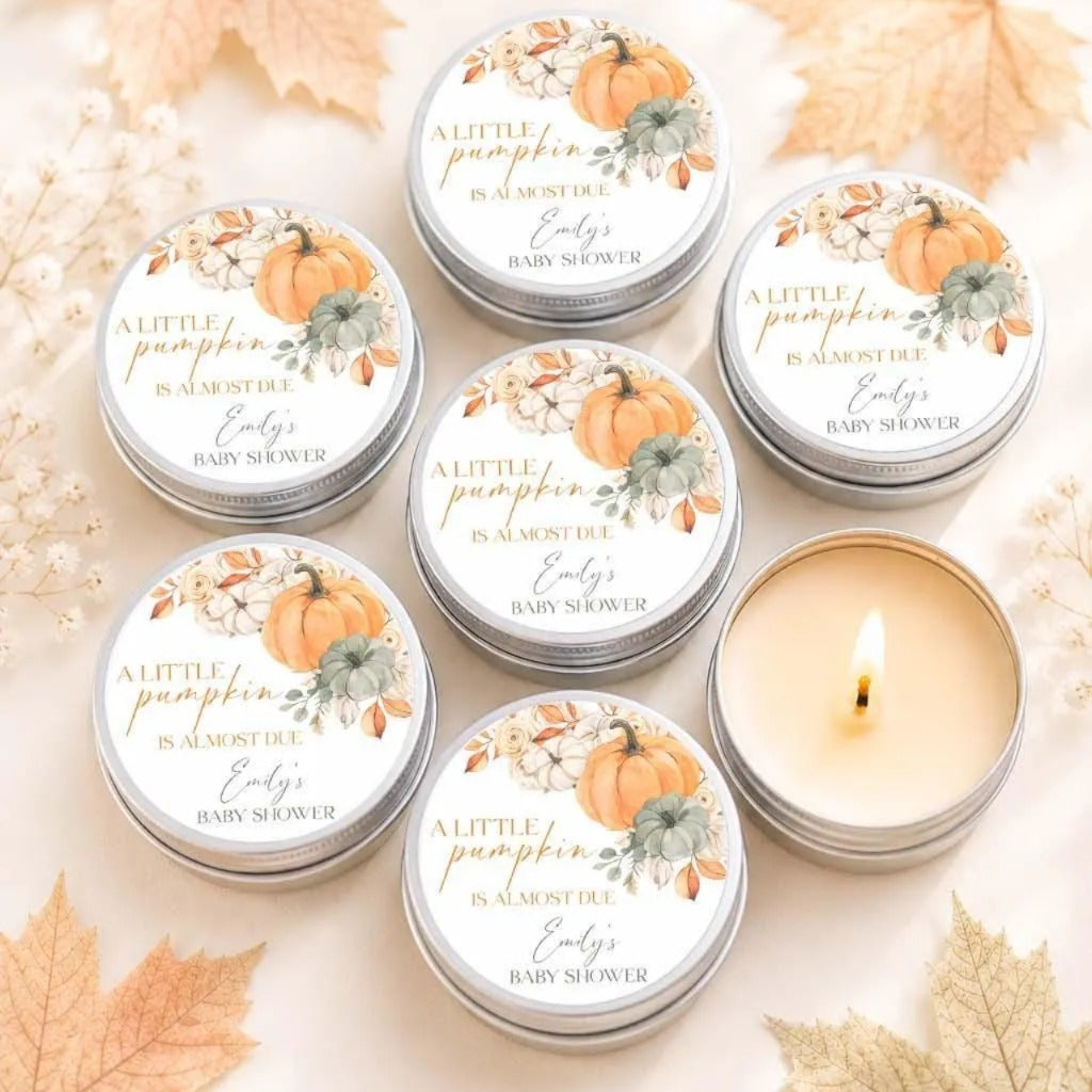 six silver tins with a pumpkin design, arranged in two rows of three. Each tin has a label that reads "A little pumpkin is almost due. Emily's baby shower". The tins are surrounded by autumn leaves, creating a festive and seasonal atmosphere.