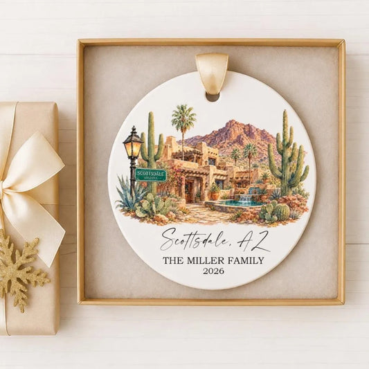 A decorative ornament featuring a desert landscape with a building, palm trees, and a mountain in the background. The ornament is displayed in a gold frame and is placed on a white surface.