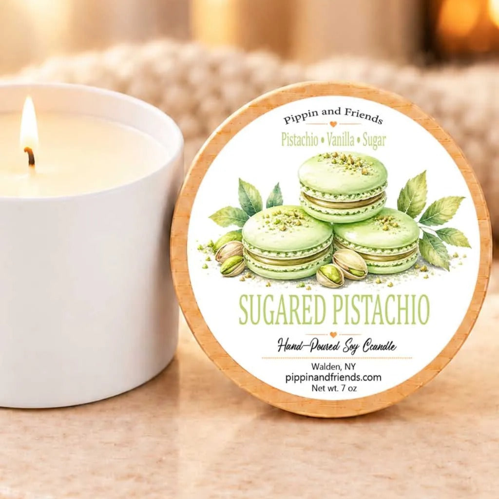 A white candle with a label that reads "SUGARED PISTACHIO" next to a lit candle on a table.