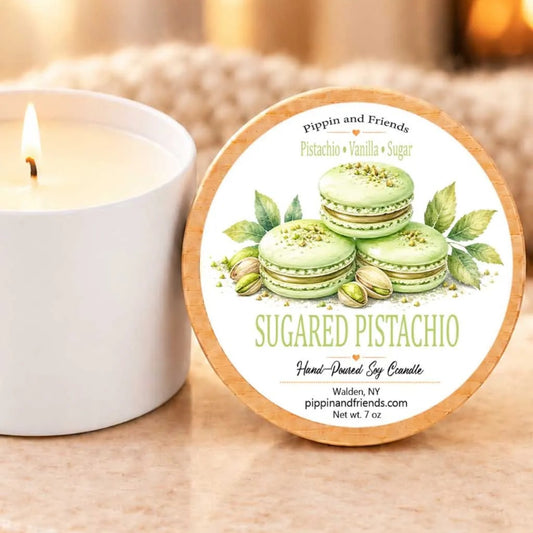 A white candle with a label that reads "SUGARED PISTACHIO" next to a lit candle on a table.