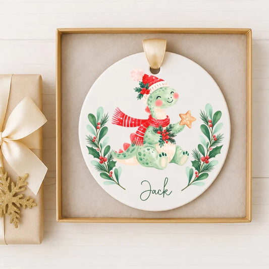 A ceramic ornament featuring a green dinosaur wearing a red scarf and hat, holding a star, and surrounded by a wreath of greenery and berries. The name "Jack" is written below the ornament.